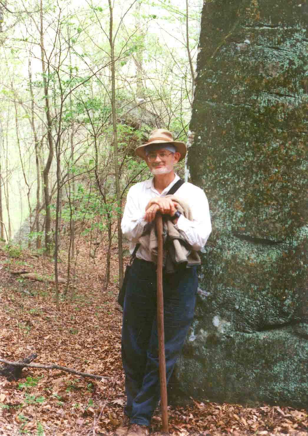 David King in natural habitat, standing with walking stick beside a tall rock.